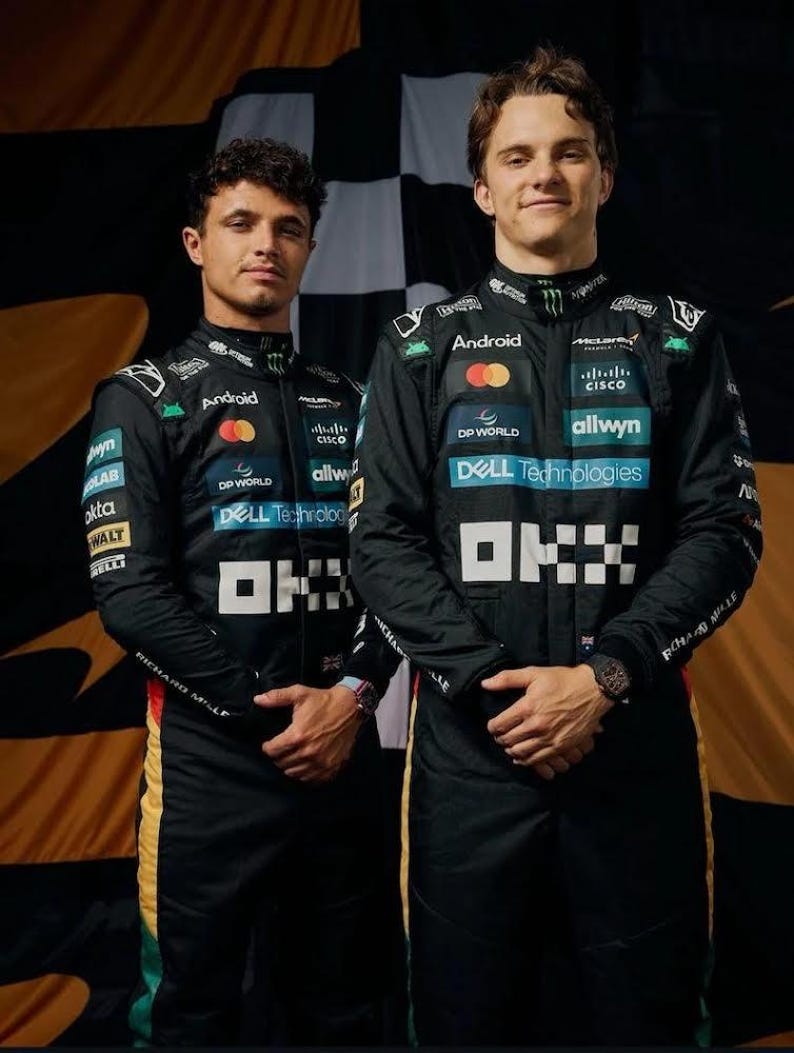 May include: Two people in black racing suits with sponsor logos, including "Android," "Dell Technologies," and "Cisco." The suits have white and yellow accents. They are standing in front of a checkered flag background.