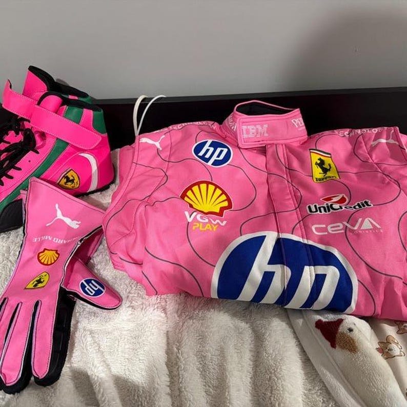 May include: Pink racing suit, gloves, and shoes with various sponsor logos, including HP, Shell, and Ferrari. The gloves have black accents and the shoes have green and black details. The suit has the IBM logo on the collar.