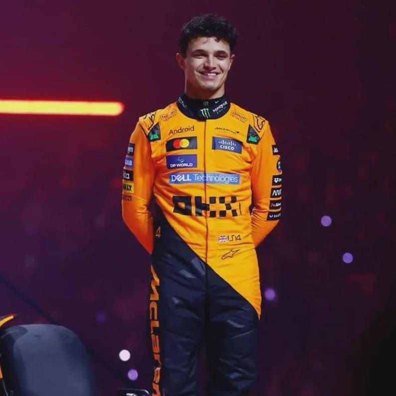 May include: A smiling person wearing a bright orange and black racing suit. The suit has multiple sponsor logos, including "Android", "Dell Technologies", and "Cisco". The suit also features the McLaren logo.
