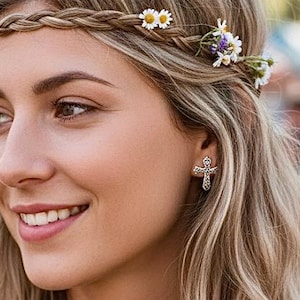 May include: A woman with long, wavy blonde hair wearing a braided headband adorned with small white and purple flowers. She is smiling, and wearing a patterned top and a cross earring.