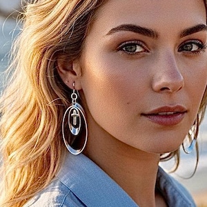 May include: A woman wearing silver oval drop earrings with a geometric design. She is wearing a light blue button-down shirt. The background features a beach with ocean waves and green foliage.