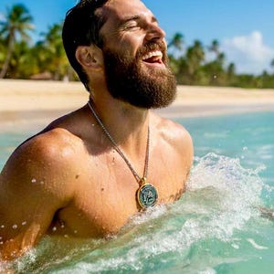 May include: A man in the ocean, smiling with his eyes closed. He is wearing a silver necklace with a round pendant. The water is turquoise, and a sandy beach with palm trees is in the background.