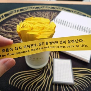 May include: A hand holds a yellow strip of paper with text in Korean and English: "The flow resumes. What seemed over comes back to life." A small cup is filled with yellow paper strips. A spiral notebook and a small notepad are in the background.