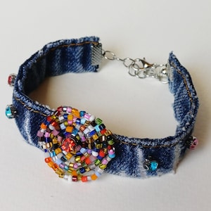 Könnte beinhalten: Ein Jeans-Armband mit einem zentralen Perlenmuster in Spiralform. Das Armband verfügt über eine Vielzahl farbiger Perlen, darunter Rot, Orange, Gelb, Grün, Blau und Lila. Kleine, farbige Edelsteine sind auf dem Jeansband angebracht.