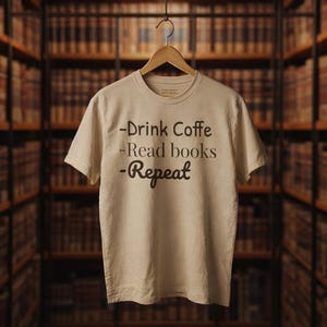 以下が含まれることがあります： 「-Drink Coffe -Read books -Repeat」の文字がダークブラウンでプリントされたベージュ色のTシャツ。木製のハンガーに掛けられており、背景には本棚が写っています。