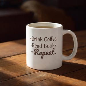 以下が含まれることがあります： 木製のテーブルの上に置かれた、コーヒーが入った白いセラミックマグカップ。マグカップには、手書き風の暗いフォントで「-Drink Coffee. -Read Books. -Repeat.」と書かれています。暖かい日差しが差し込んでいます。