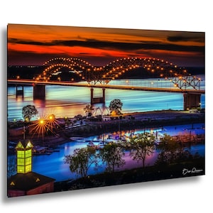 May include: A vibrant photograph of a bridge at dusk, with the sky ablaze in orange and red hues. The bridge is illuminated with warm lights, reflecting in the blue water below. Trees and buildings line the riverbank.