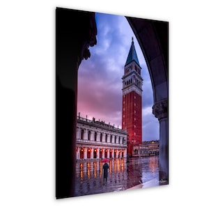 Peut inclure: Photographie de la Campanile de Saint-Marc à Venise, Italie, encadrée par une arche. La tour en briques rouges se dresse contre un ciel dramatique et coloré. Une personne avec un parapluie rouge marche sur la place mouillée.