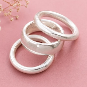 May include: Three polished silver rings with smooth, rounded bands. The rings are stacked in an overlapping arrangement, showcasing a bright, reflective metallic finish. Each band features a consistent, simple circular shape with no additional embellishments.