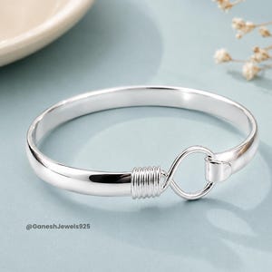 May include: A polished silver bangle bracelet featuring a smooth, rounded band that connects with a coiled wire detail and a small circular loop clasp. The text @GaneshJewels925 is printed in the lower left corner of the image.