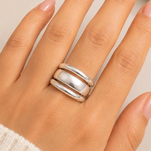 Puede incluir: Un anillo de tono plateado que presenta tres bandas distintas, lisas y redondeadas, apiladas juntas. El diseño consta de dos bandas exteriores más delgadas y una banda central ligeramente más ancha y gruesa, con un acabado metálico pulido.