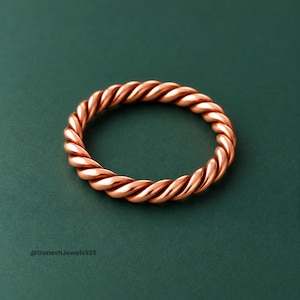 Puede incluir: Un anillo de cobre pulido que presenta un diseño de cuerda trenzada continua. El metal tiene un tono marrón rojizo metálico cálido con contornos suaves y redondeados en toda la banda. El texto GaneshJewels925 es visible en la esquina inferior izquierda.