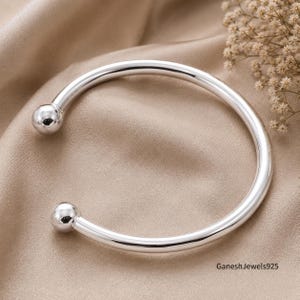 May include: A polished silver cuff bracelet featuring a smooth, circular band that terminates in two spherical ball ends. The metal surface is highly reflective and bright. The text GaneshJewels925 is printed in the bottom right corner.
