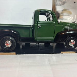 May include: A die-cast model of a vintage green pickup truck with black fenders and wheels. The truck has a detailed cab, bed, and chrome accents. The wheels have red centre caps.