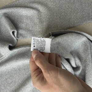 Op de afbeelding: Een grijze trui met een zwarte streep. De trui is gemaakt van een zacht materiaal en heeft een etiket met wasvoorschriften. De trui is opgevouwen en het etiket wordt door een hand vastgehouden.