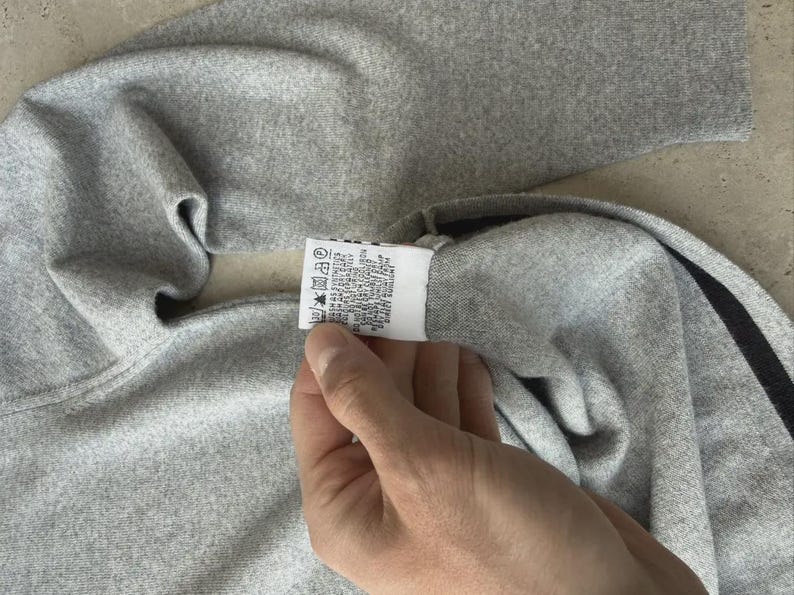 Op de afbeelding: Een grijze trui met een zwarte streep. De trui is gemaakt van een zacht materiaal en heeft een etiket met wasvoorschriften. De trui is opgevouwen en het etiket wordt door een hand vastgehouden.