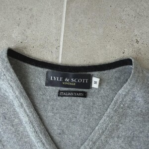 Op de afbeelding: Een grijze V-hals trui met een zwarte rand rond de halslijn. Het label luidt "LYLE & SCOTT VINTAGE" en "ITALIAN YARN". De trui is maat medium.