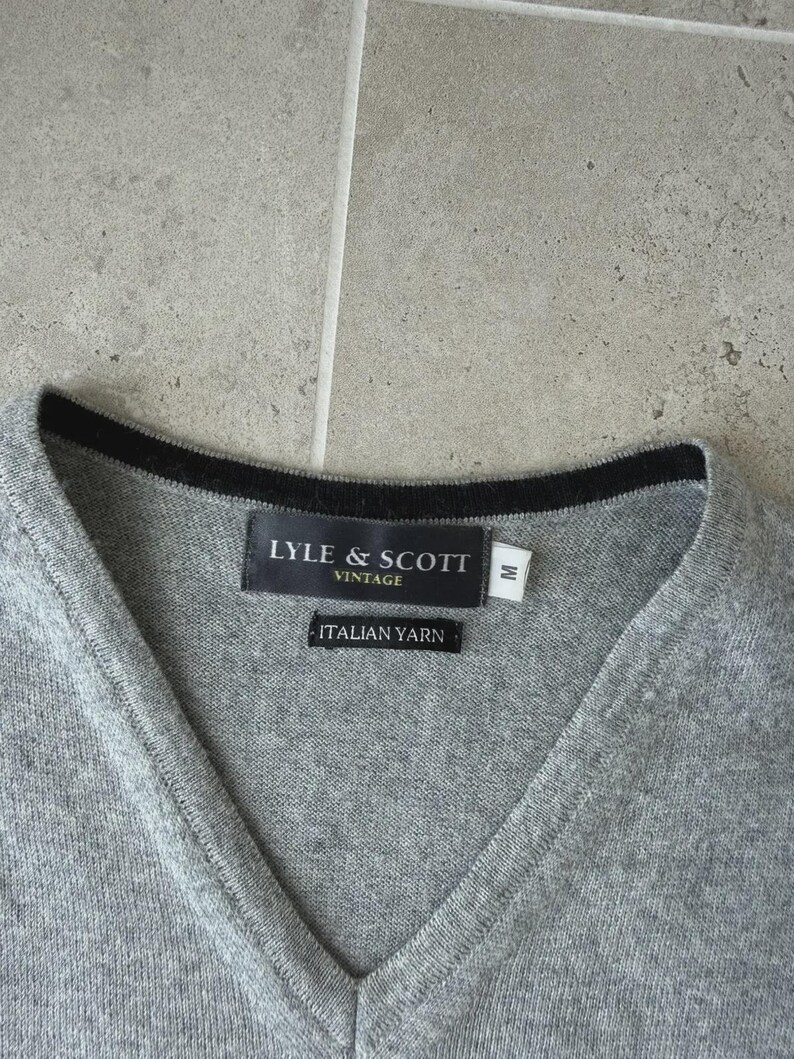 Op de afbeelding: Een grijze V-hals trui met een zwarte rand rond de halslijn. Het label luidt "LYLE & SCOTT VINTAGE" en "ITALIAN YARN". De trui is maat medium.