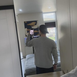 Op de afbeelding: Een persoon met een grijs shirt met lange mouwen en een zwarte baseballcap met een Nike-logo, weerspiegeld in een grote spiegel. De persoon houdt een telefoon vast. De achtergrond omvat een bed, een schilderij en een raam.