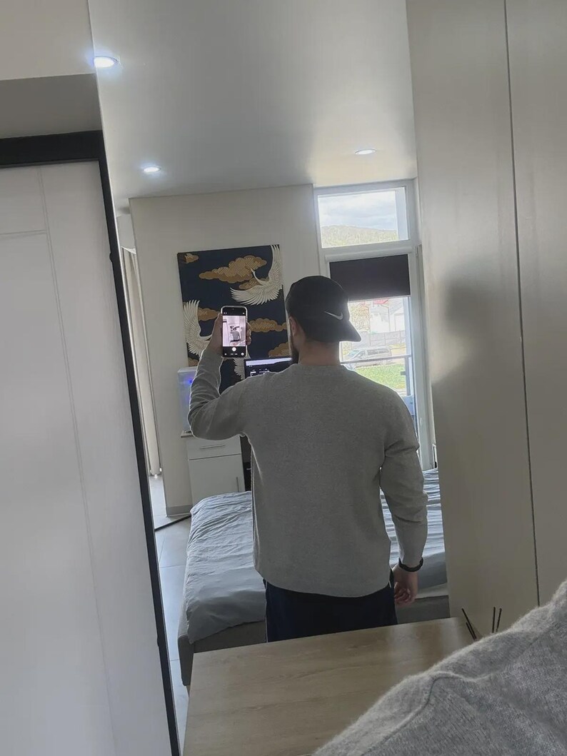 Op de afbeelding: Een persoon met een grijs shirt met lange mouwen en een zwarte baseballcap met een Nike-logo, weerspiegeld in een grote spiegel. De persoon houdt een telefoon vast. De achtergrond omvat een bed, een schilderij en een raam.