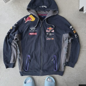 Puede incluir: Sudadera con capucha azul marino con cremallera, capucha y varios logotipos, incluyendo "Red Bull" e "Infiniti". La chaqueta tiene paneles laterales grises y detalles de cremallera morados. La capucha tiene el texto "Pepe Jeans" impreso.