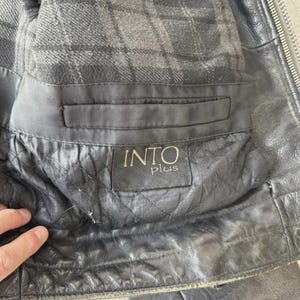 May include: Close-up of a black leather jacket interior. The lining is a gray plaid pattern. The jacket has a pocket with a label that reads "INTO plus". The leather has a quilted design.