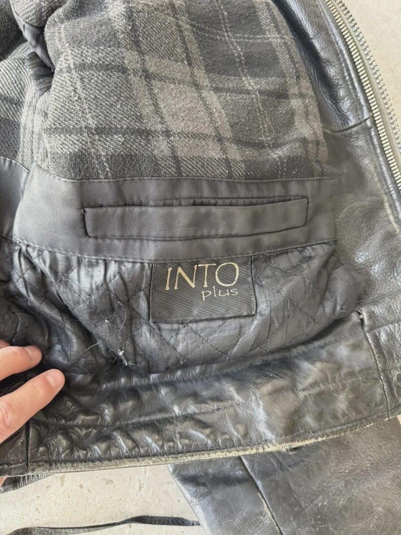 May include: Close-up of a black leather jacket interior. The lining is a gray plaid pattern. The jacket has a pocket with a label that reads "INTO plus". The leather has a quilted design.