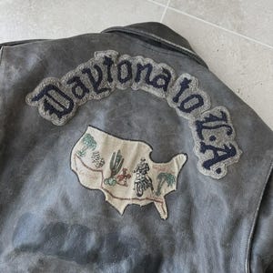 May include: A gray leather jacket with embroidered patches. The patches include the text "Daytona to L.A." in a curved design, a map of the U.S. with a motorcycle rider, and the number "93".