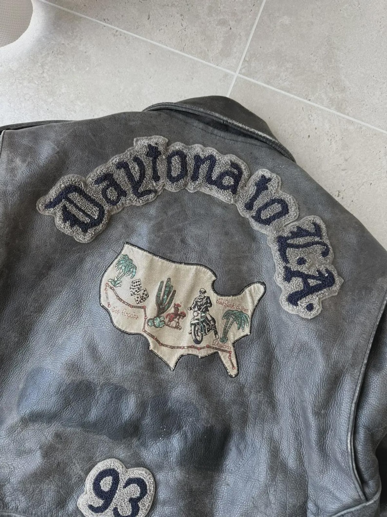 May include: A gray leather jacket with embroidered patches. The patches include the text "Daytona to L.A." in a curved design, a map of the U.S. with a motorcycle rider, and the number "93".