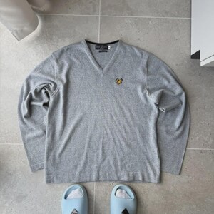 Op de afbeelding: Een lichtgrijs V-hals shirt met lange mouwen en een klein geel embleem op de borst. Gecombineerd met lichtblauwe slippers en witte sokken. De halslijn heeft een zwarte rand. Het merk is zichtbaar.
