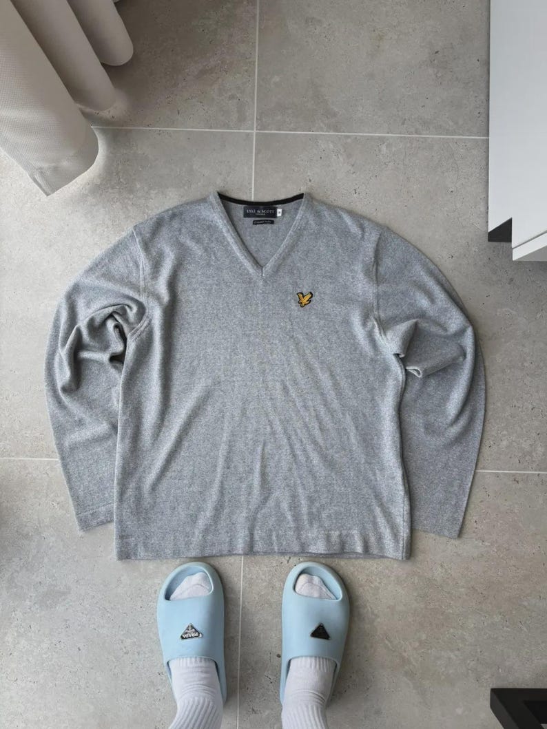 Op de afbeelding: Een lichtgrijs V-hals shirt met lange mouwen en een klein geel embleem op de borst. Gecombineerd met lichtblauwe slippers en witte sokken. De halslijn heeft een zwarte rand. Het merk is zichtbaar.