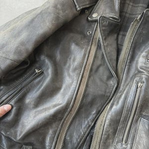 May include: A worn, dark gray leather jacket with multiple zippered pockets and a notched collar. The jacket has a distressed appearance, with visible wear and tear on the leather. The interior lining features a plaid pattern.