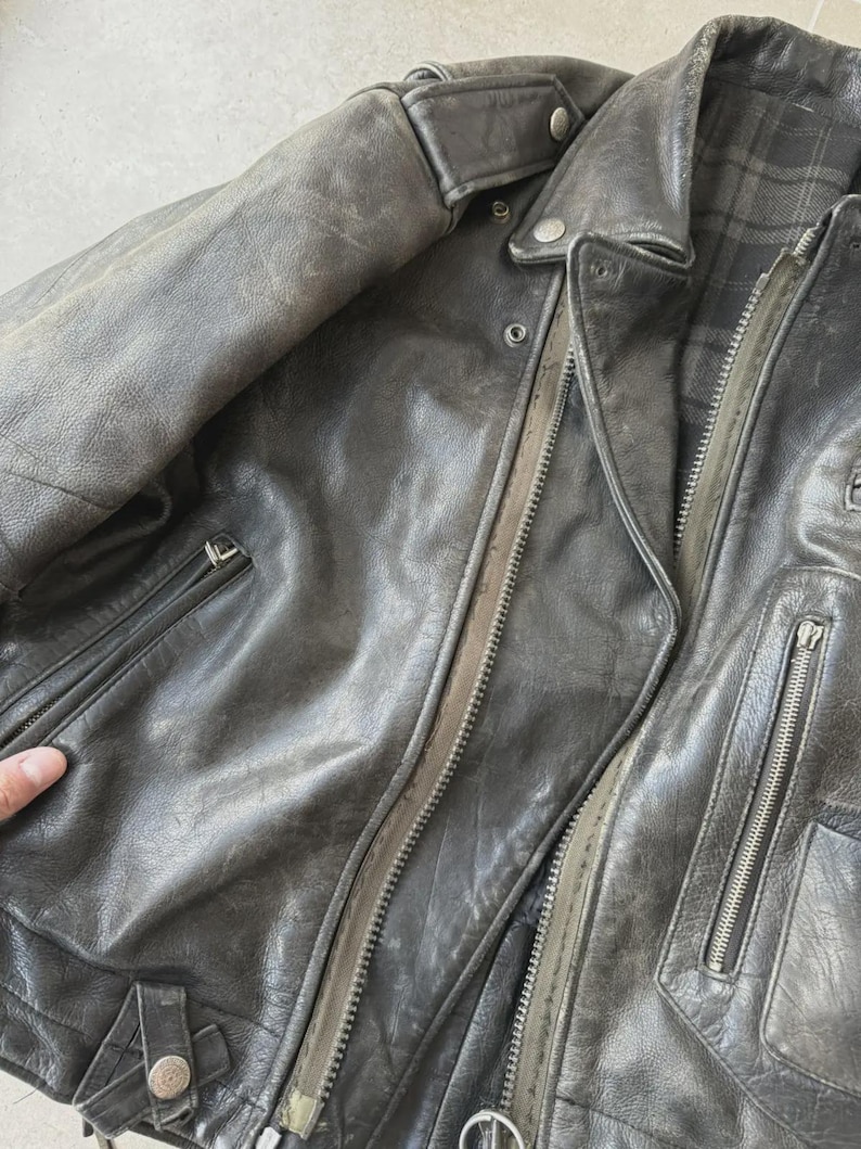 May include: A worn, dark gray leather jacket with multiple zippered pockets and a notched collar. The jacket has a distressed appearance, with visible wear and tear on the leather. The interior lining features a plaid pattern.