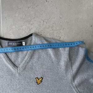 Op de afbeelding: Een grijze V-hals trui met zwarte bies en een geel vogel logo. De trui wordt gemeten met een blauwe meetlint. De merknaam "LYLE & SCOTT" is zichtbaar. De trui is ongeveer 46 cm breed.