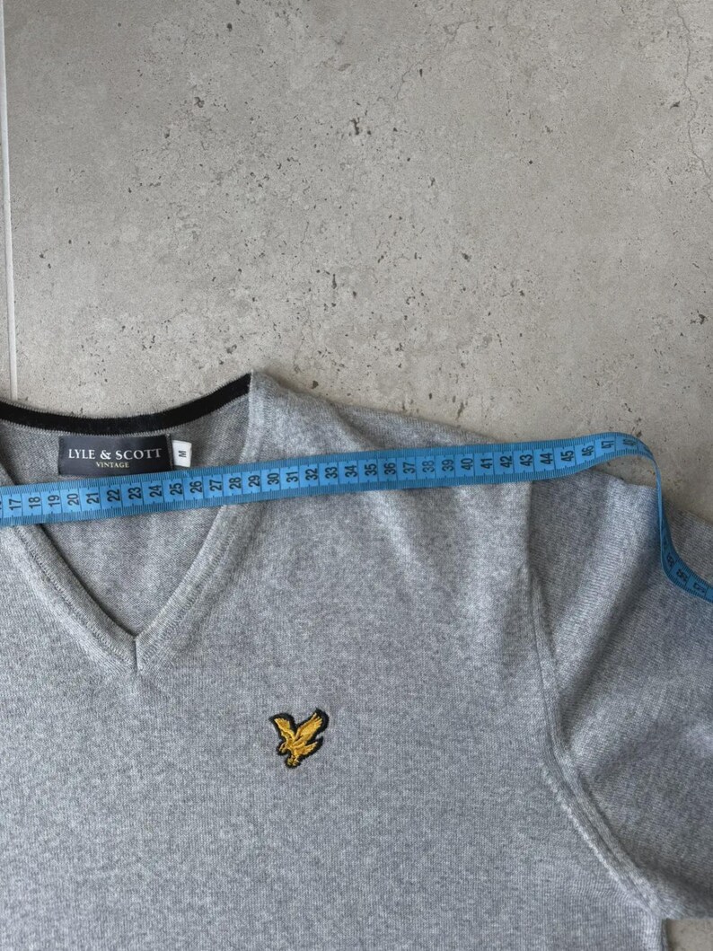 Op de afbeelding: Een grijze V-hals trui met zwarte bies en een geel vogel logo. De trui wordt gemeten met een blauwe meetlint. De merknaam "LYLE & SCOTT" is zichtbaar. De trui is ongeveer 46 cm breed.