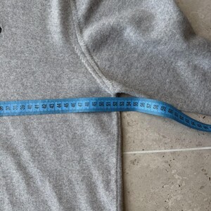 Op de afbeelding: Een lichtgrijze trui met een blauwe meetlint die een lengte van ongeveer 56 cm aangeeft. De trui heeft een ronde hals en lange mouwen. De stof lijkt een zachte brei te zijn.