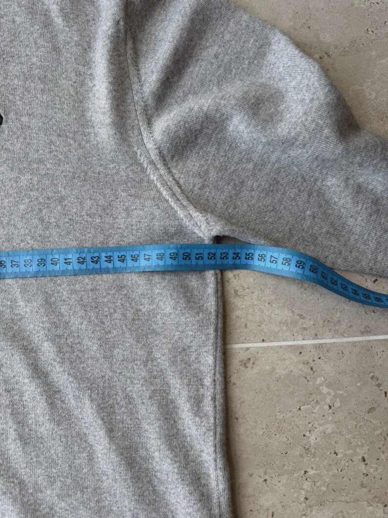 Op de afbeelding: Een lichtgrijze trui met een blauwe meetlint die een lengte van ongeveer 56 cm aangeeft. De trui heeft een ronde hals en lange mouwen. De stof lijkt een zachte brei te zijn.