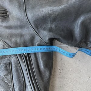 May include: A gray leather jacket with a zipper closure and multiple pockets. A blue measuring tape is laid across the jacket, showing a measurement of approximately 24 inches.