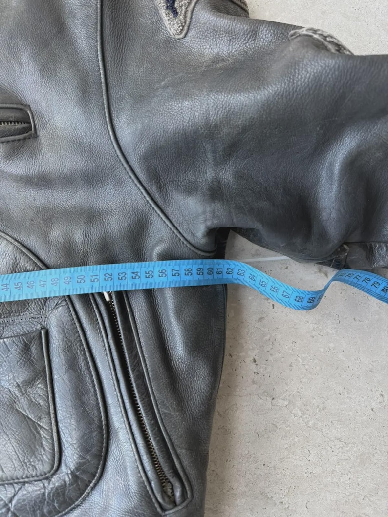 May include: A gray leather jacket with a zipper closure and multiple pockets. A blue measuring tape is laid across the jacket, showing a measurement of approximately 24 inches.