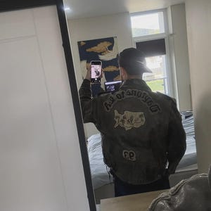 May include: A dark gray leather jacket with patches on the back. The patches include text that reads "AGE OF ANOTHER" and a map of Cyprus. The jacket is worn with a black cap.