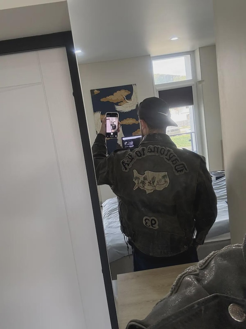 May include: A dark gray leather jacket with patches on the back. The patches include text that reads "AGE OF ANOTHER" and a map of Cyprus. The jacket is worn with a black cap.
