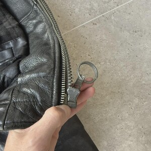 May include: Close-up of a black leather jacket with a silver zipper. The jacket has a textured appearance and a quilted lining. The zipper pull is silver and attached to a metal ring.