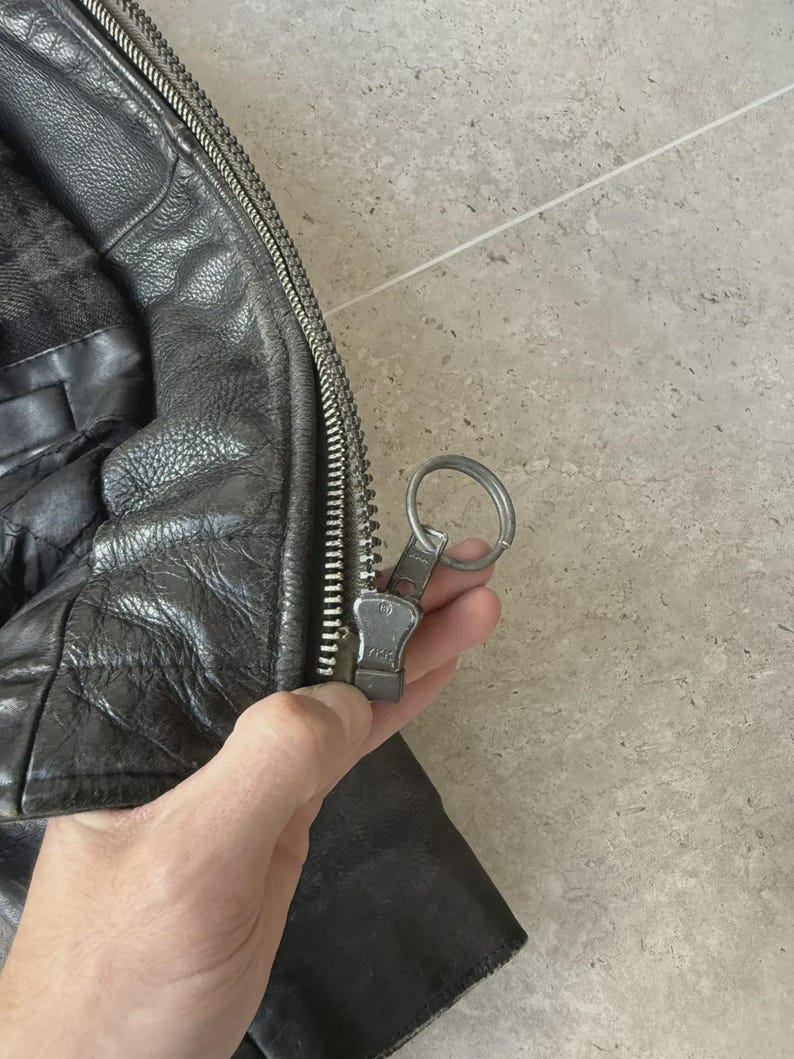 May include: Close-up of a black leather jacket with a silver zipper. The jacket has a textured appearance and a quilted lining. The zipper pull is silver and attached to a metal ring.