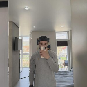 Op de afbeelding: Een persoon in een spiegel, gekleed in een grijs shirt met lange mouwen en een zwarte baseballcap. Het shirt heeft een klein geel logo. De persoon houdt een telefoon vast.