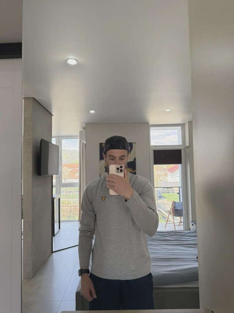 Op de afbeelding: Een persoon in een spiegel, gekleed in een grijs shirt met lange mouwen en een zwarte baseballcap. Het shirt heeft een klein geel logo. De persoon houdt een telefoon vast.