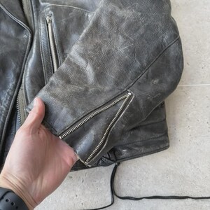 May include: A distressed, dark gray leather jacket with silver zippers on the sleeves and pockets. The jacket has a worn, vintage appearance, with visible wear and tear on the leather. A patch is visible on the shoulder.