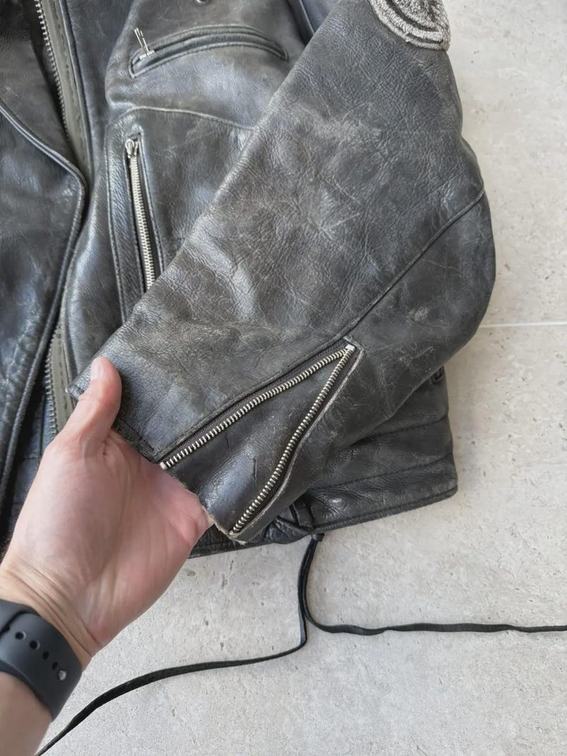 May include: A distressed, dark gray leather jacket with silver zippers on the sleeves and pockets. The jacket has a worn, vintage appearance, with visible wear and tear on the leather. A patch is visible on the shoulder.