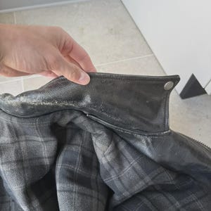 May include: Close-up of a black leather jacket with a plaid interior. The jacket has a classic collar and a button detail. The interior lining features a gray and black plaid pattern. The jacket appears to be well-worn.