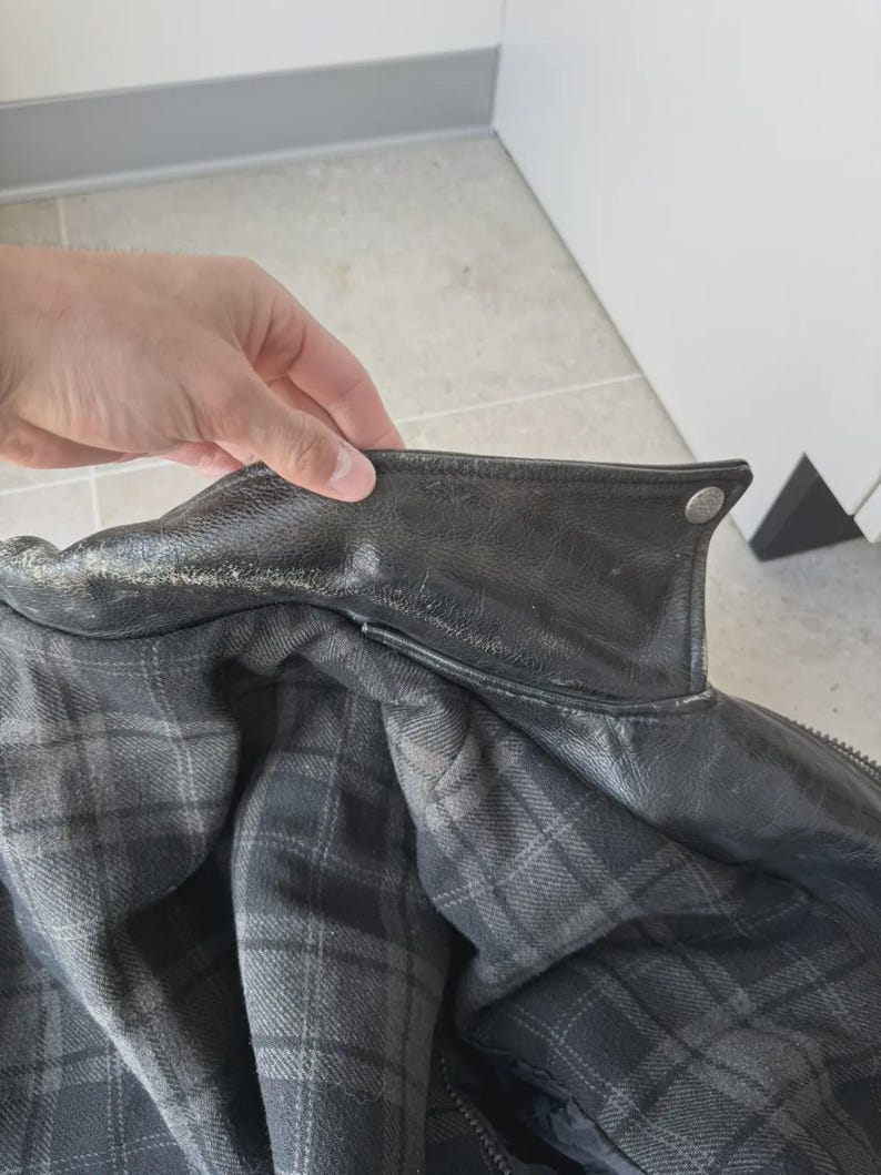 May include: Close-up of a black leather jacket with a plaid interior. The jacket has a classic collar and a button detail. The interior lining features a gray and black plaid pattern. The jacket appears to be well-worn.