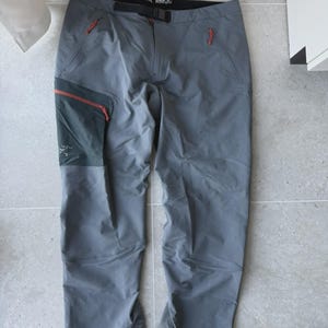 Könnte beinhalten: Graue Outdoor-Hose mit roten Reißverschlussakzenten und einem schwarzen Gürtel. Die Hose hat mehrere Taschen und verstärkte Einsätze. Hergestellt aus strapazierfähigem, wasserabweisendem Material, geeignet für Outdoor-Aktivitäten.